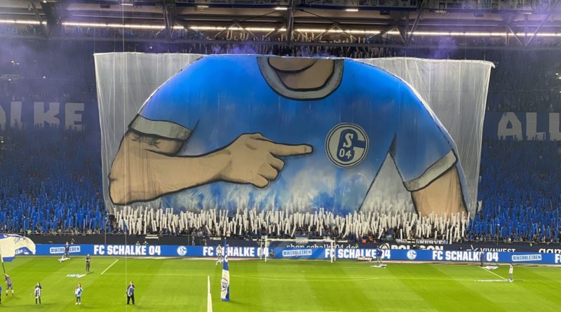 Schalker Choreografie am 30. Oktober zum Heimspiel gegen Freiburg. Vor der Nordkurve wird das Bild eines Spielers hochgeogen, das sich langsam mit blauem Rauch füllt und es so blau färbt.