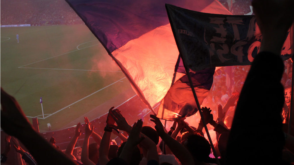 Fans im Stadion, hinter Fahnen brennt eine bengalische Fackel und beleuchtet diese von hinten
