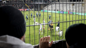 Fans hinter einem Zaun, gucken auf das Spielfeld, wo ein Schalke-Spiel stattfindet